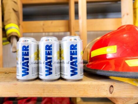 Three cans of emergency drinking water are positioned on a wooden workbench next to a red and yellow fireman's helmet.